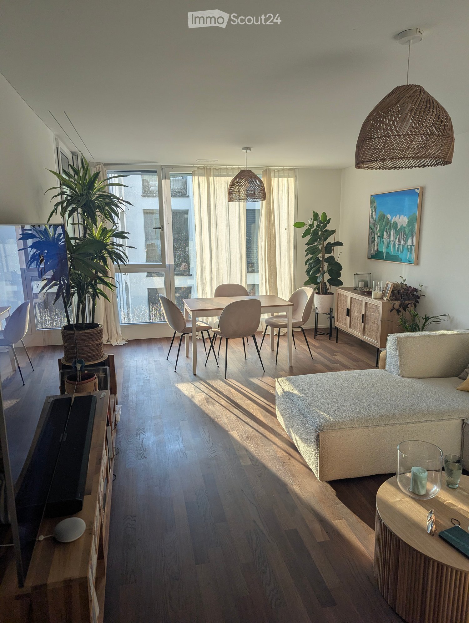A modern, well-lit living space with a sofa, dining table, chairs, cabinet, potted plants, and artwork. The room features wooden flooring and large windows with sheer curtains.