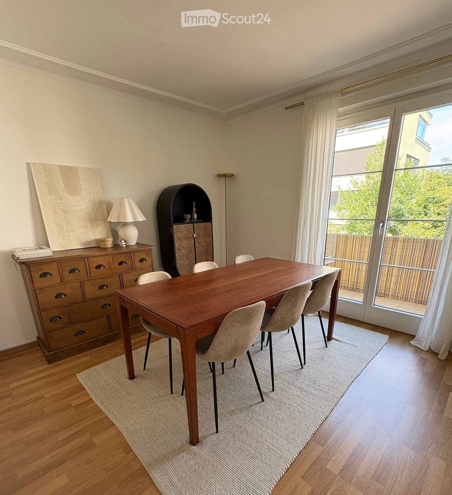 Dining room with a wooden dining table, six chairs, a wooden cabinet, a lamp, and a large picture frame on the wall. The room has large windows with white curtains and a view of a garden.