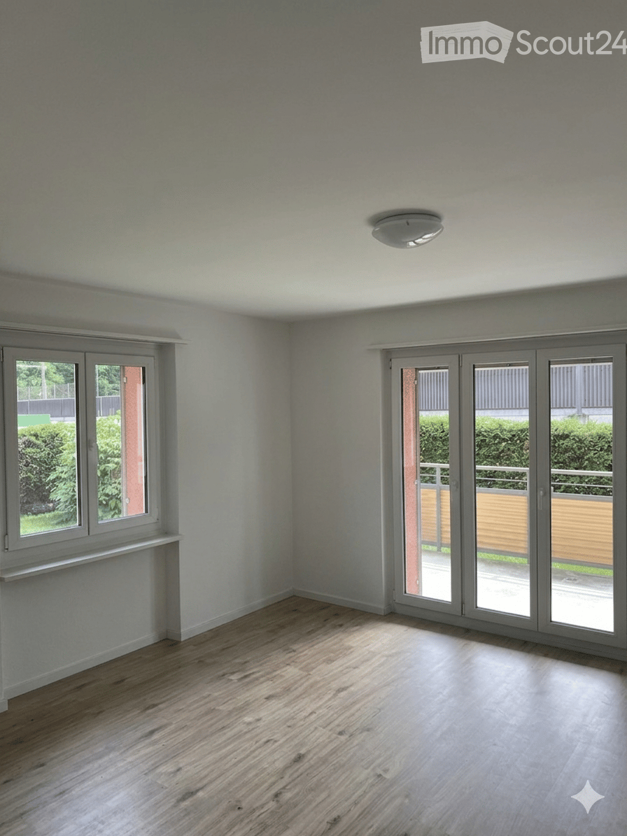 Empty room with light grey walls, wooden flooring, large window, and sliding glass doors leading to a balcony.