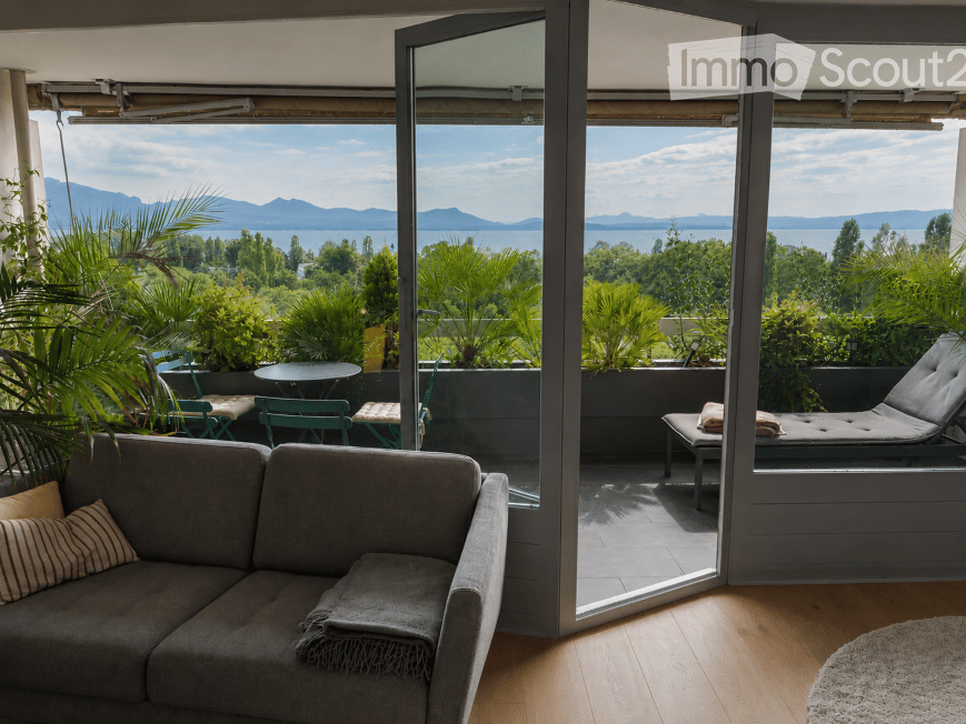 Living room with a grey sofa, wooden floor, large windows opening to a balcony with outdoor furniture and scenic mountain view.