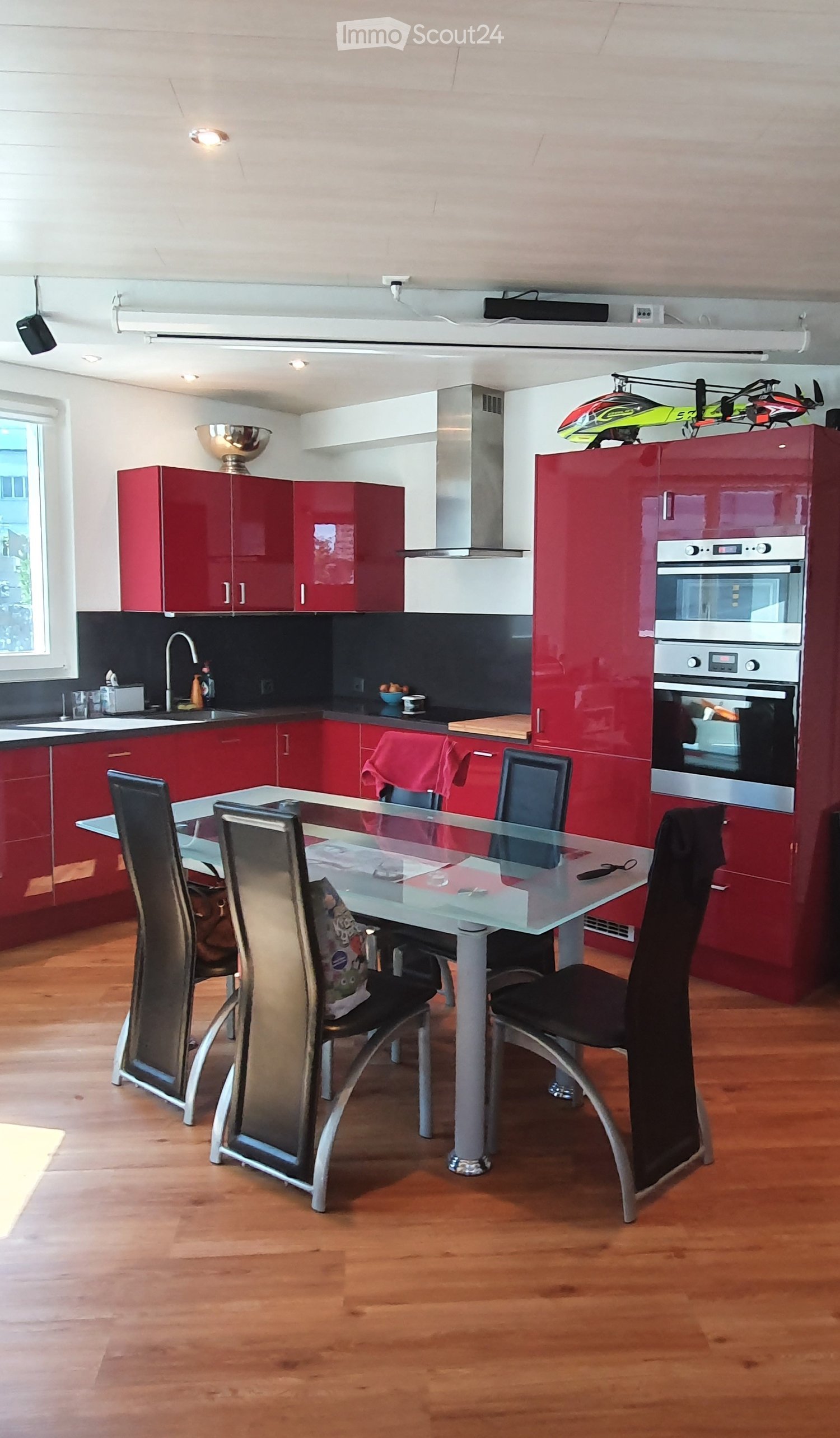 Modern kitchen with red cabinets, stainless steel appliances, a glass dining table with four chairs, and a wooden floor.