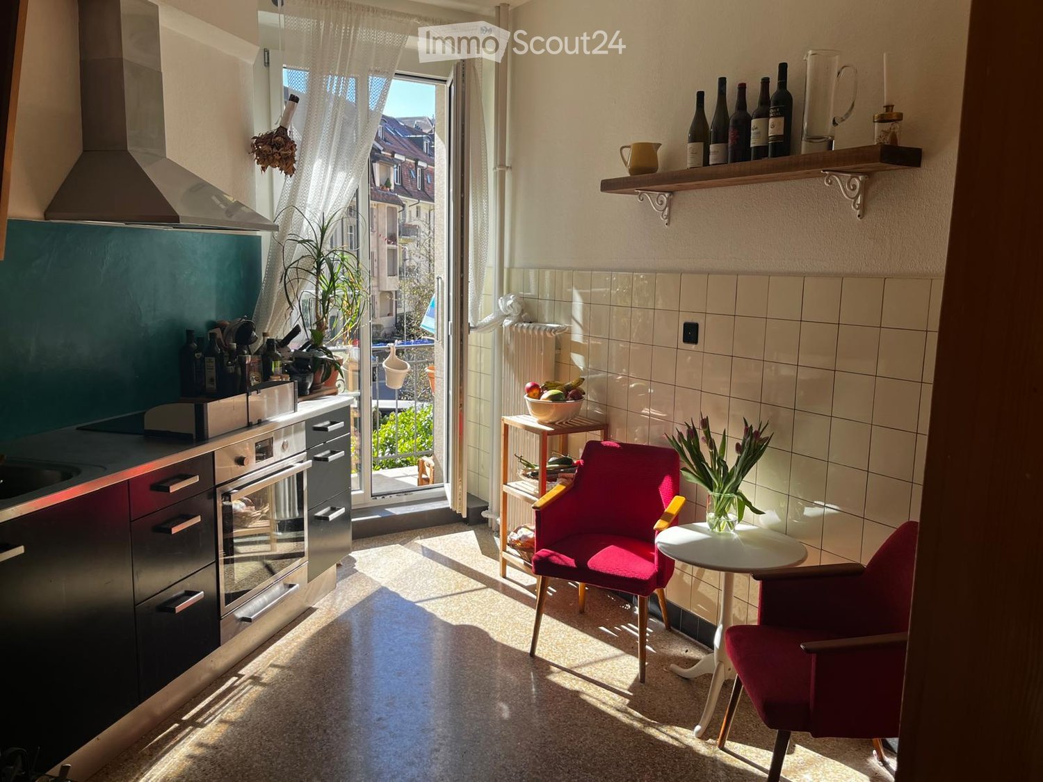 A small, well-lit kitchen with modern appliances, a red chair, a small table, and shelves with bottles and a vase.