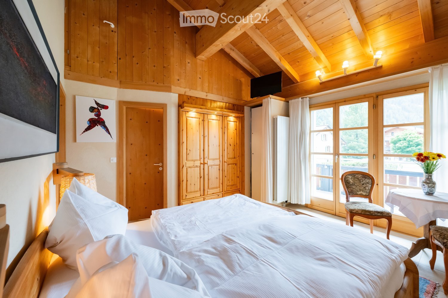 Cozy bedroom with wooden beams, a large bed with white linens, a wooden wardrobe, a small table with a vase of flowers, and a chair near the window.