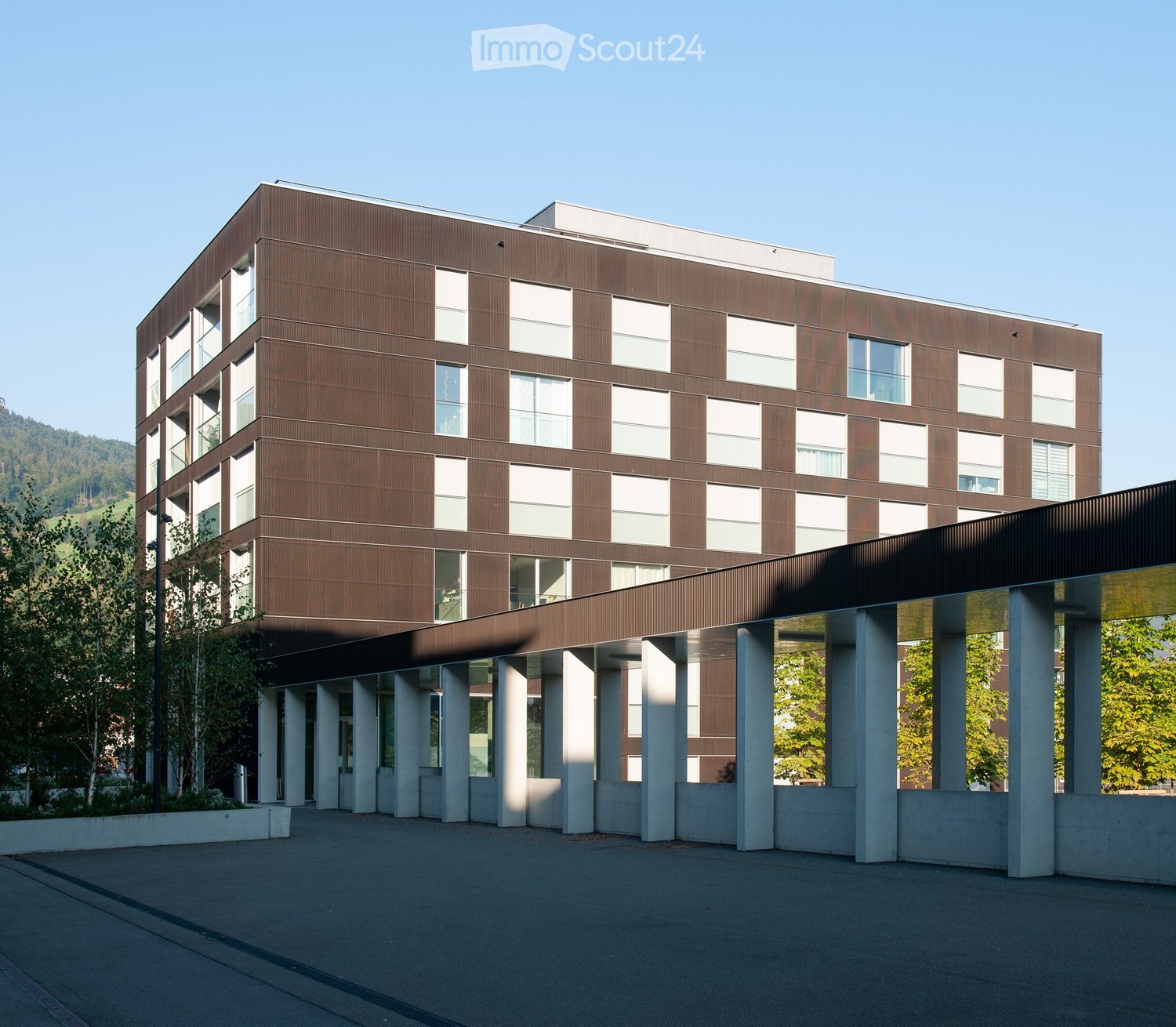 Modern multi-story building with brown and white facade, large windows, and a covered entrance supported by concrete columns.