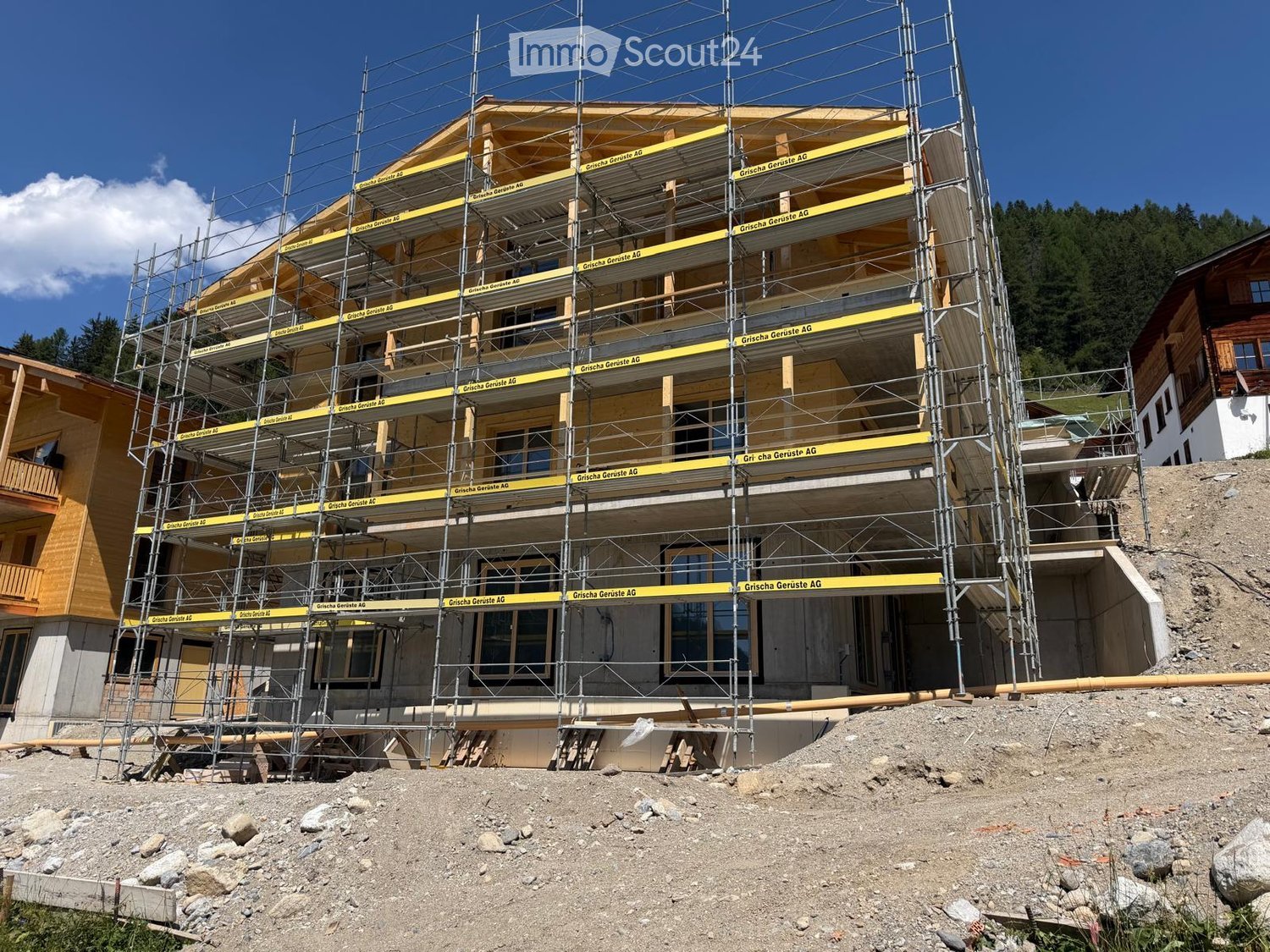 A multi-story building under construction, covered in scaffolding with exposed wooden framing and yellow safety tape. The structure is situated on a raised ground floor with visible construction materials and debris around the base.