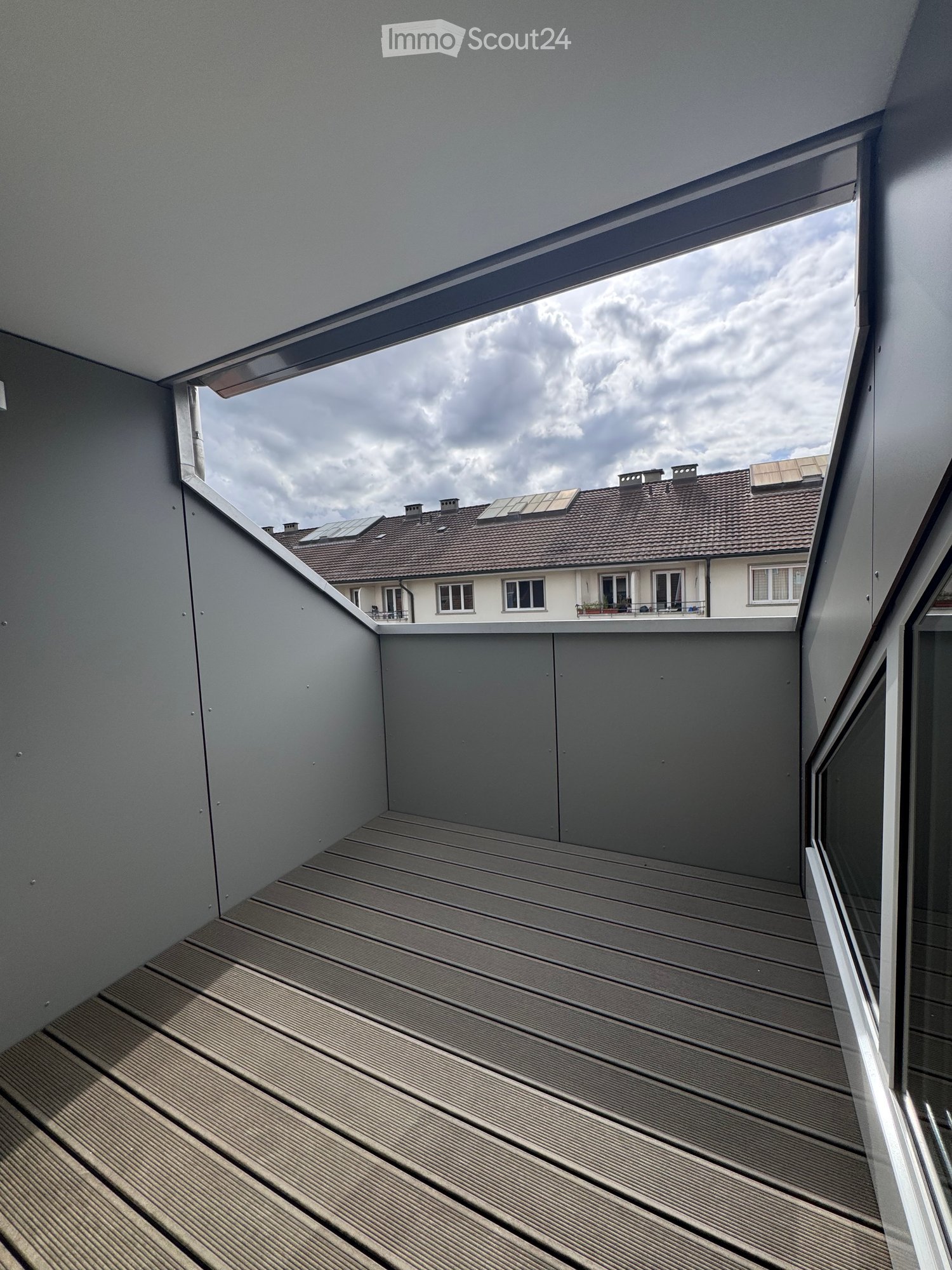 Modern wooden deck balcony with grey walls, large window offering a view of neighboring rooftops and solar panels.
