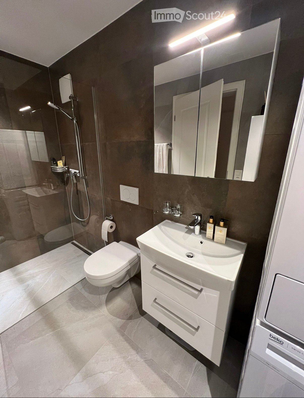 Modern bathroom with dark brown walls, white tiles, a toilet, a sink with a mirror, a shower area, and a towel rack.