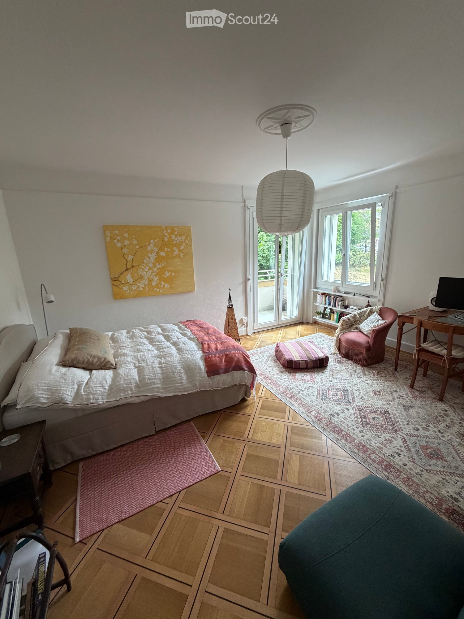 Cozy bedroom with a bed, sofa, chair, small table with laptop, bookshelf, and decorative rug. Features large windows and a sliding glass door leading to a balcony.