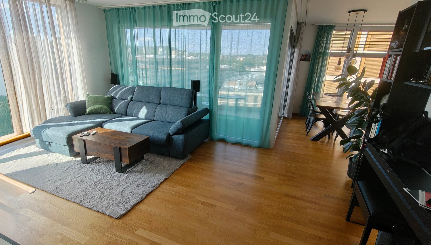 Modern living room with a grey sectional sofa, wooden coffee table, dining area with chairs and table, bookshelf, and a potted plant. Large windows with sheer curtains let in natural light.