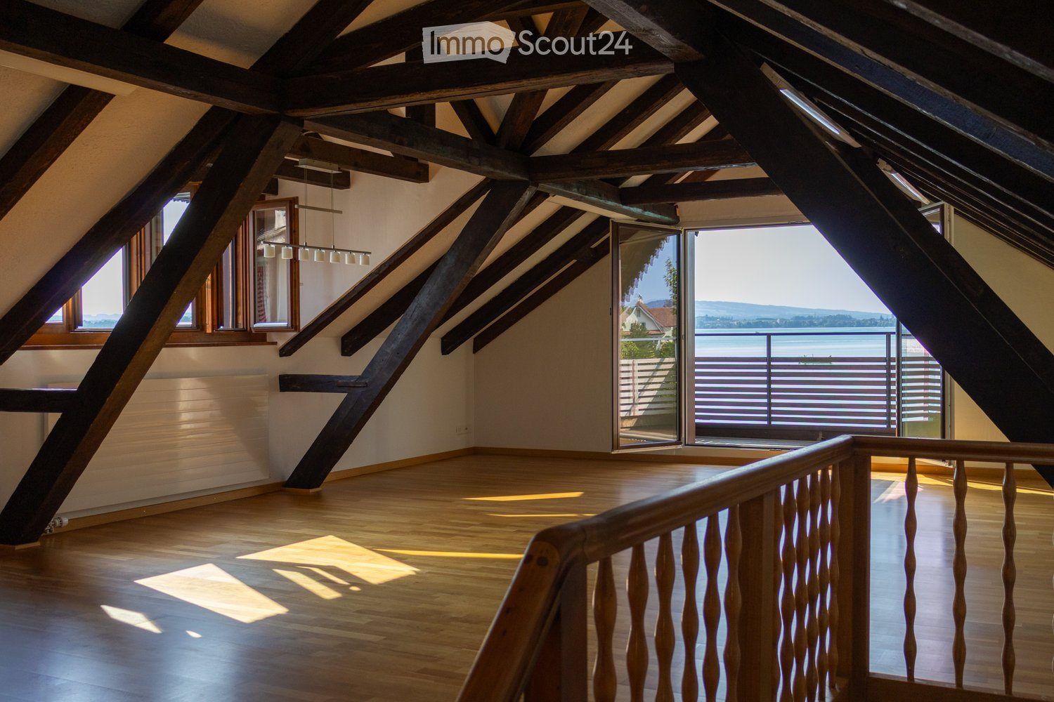 An attic room with wooden beams, wooden flooring, and large windows offering a scenic view of the lake and surrounding landscape.
