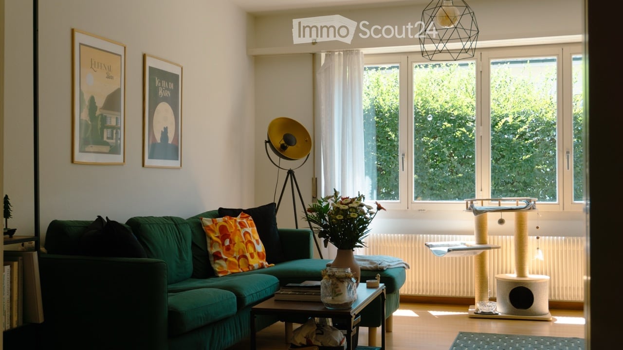 A modern living room with a green sofa, colorful throw pillow, coffee table with books and a vase of flowers, cat tree and cat bed, large windows with white curtains, and decorative wall art.