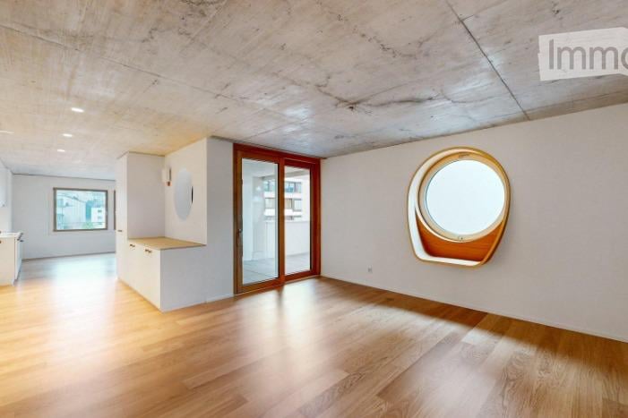 Empty room with wooden flooring, white walls, and large windows, including a round porthole window and a sliding glass door leading to another space.