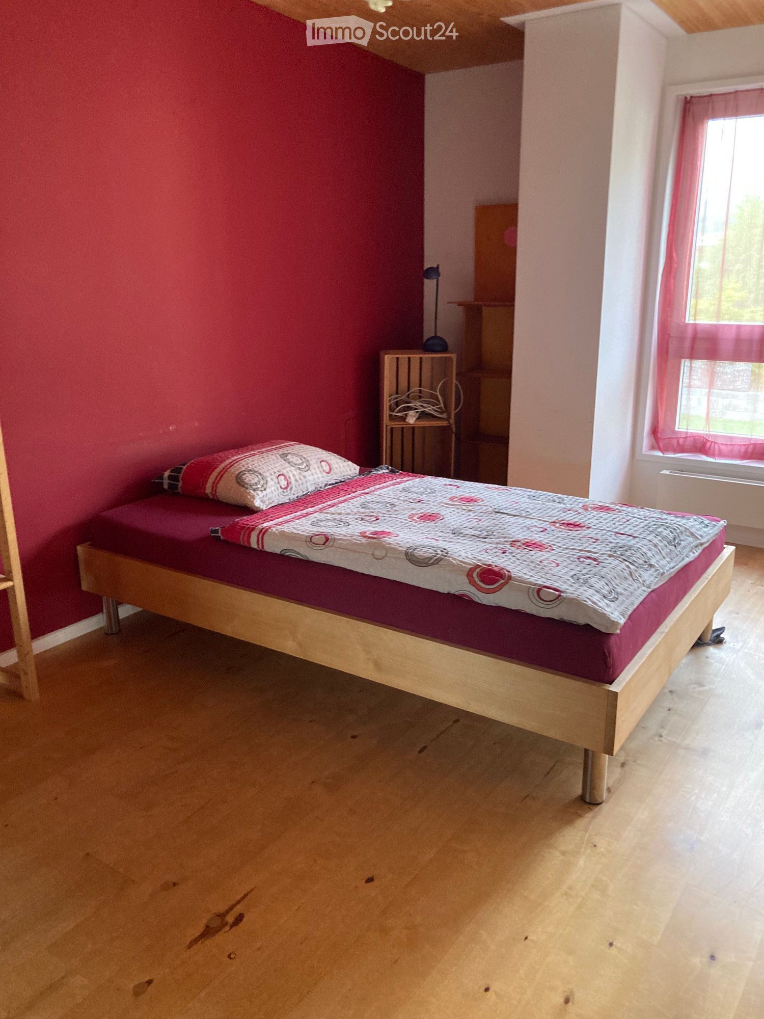 Bedroom with wooden floor, red accent wall, single bed with patterned bedding, small nightstand with lamp, window with pink curtain.