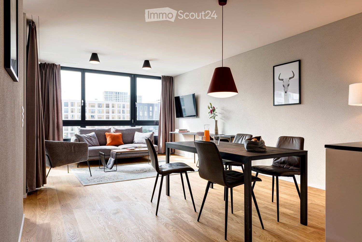 Modern living room with a grey sofa, wooden dining table, chairs, pendant light, television, and decorative items.