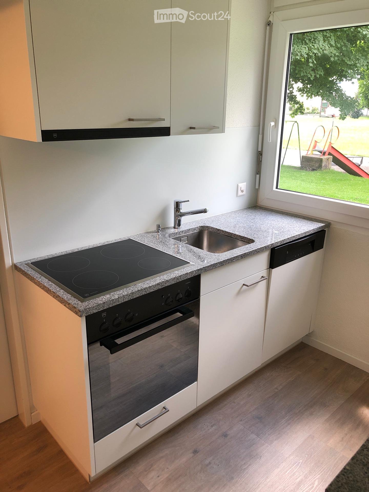 Modern kitchen with white cabinets, granite countertop, induction stove, oven, sink, and dishwasher. Large window offers view of outdoor area.