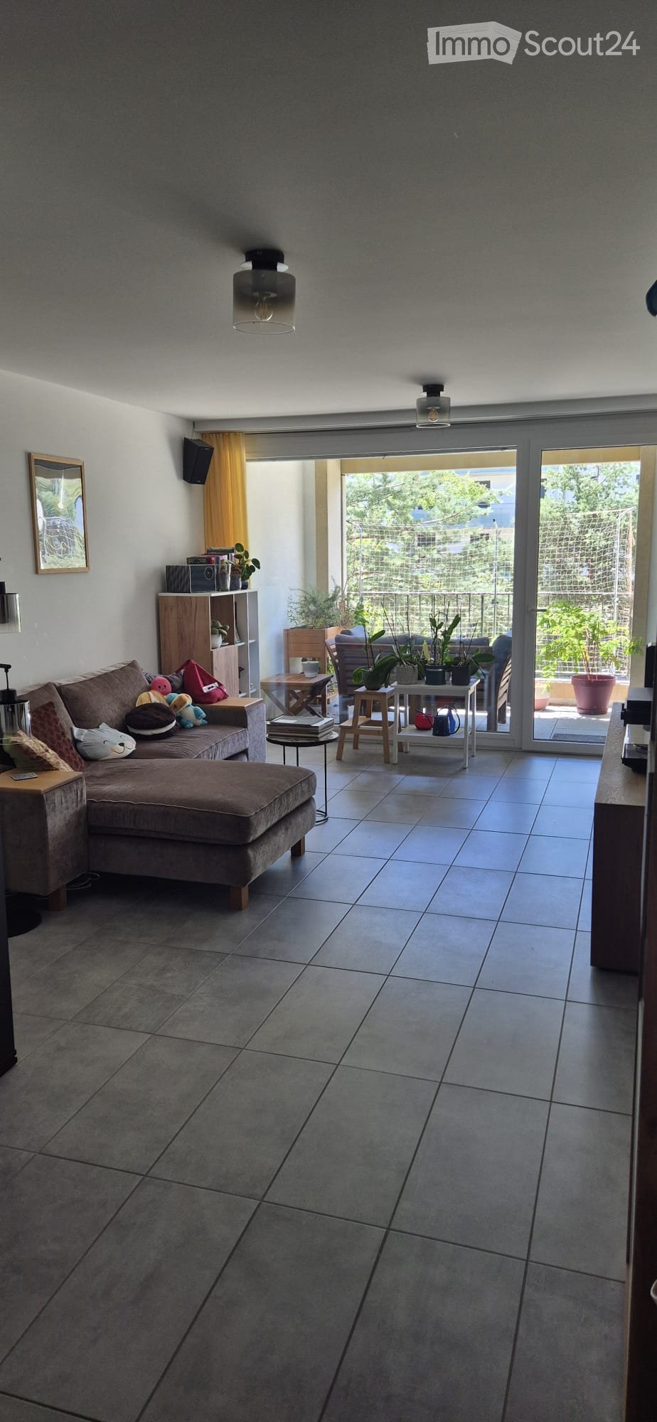 Modern living room with grey sofa, ottoman, small tables, and shelves. Large sliding glass doors open to a balcony with plants.