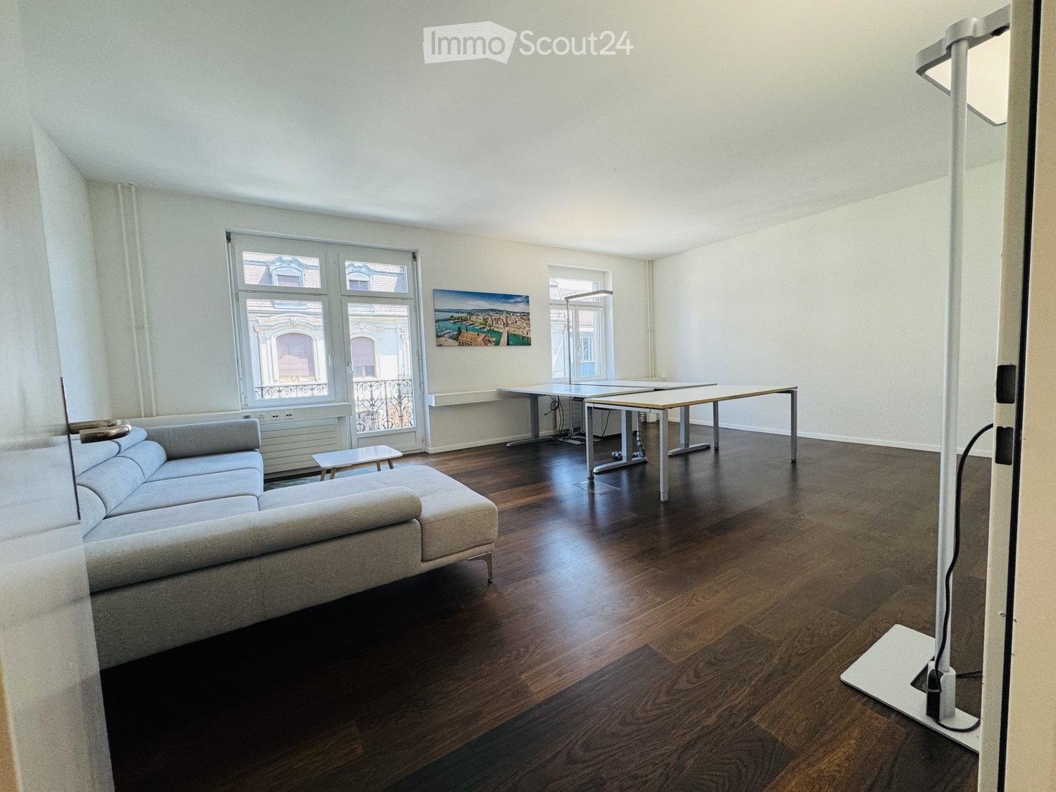 Bright living room with wooden flooring, large windows, a grey sectional sofa, a coffee table, and a dining table with chairs.