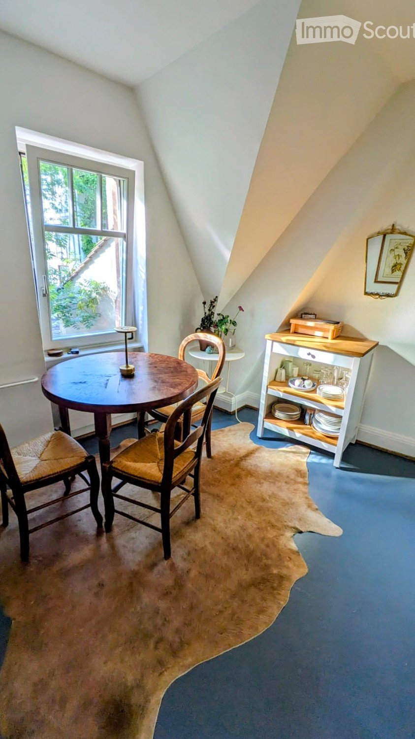 Cozy attic room with slanted ceilings, blue carpet, round wooden table, chairs, small cabinet, and decorative items.