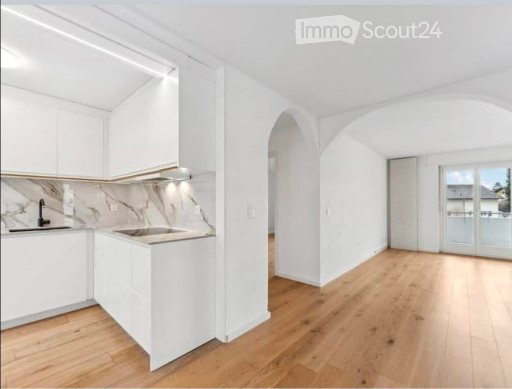 Modern kitchen with white cabinets, marble countertop, and wooden flooring, connected to a bright living space with large windows and sliding doors leading to a balcony.