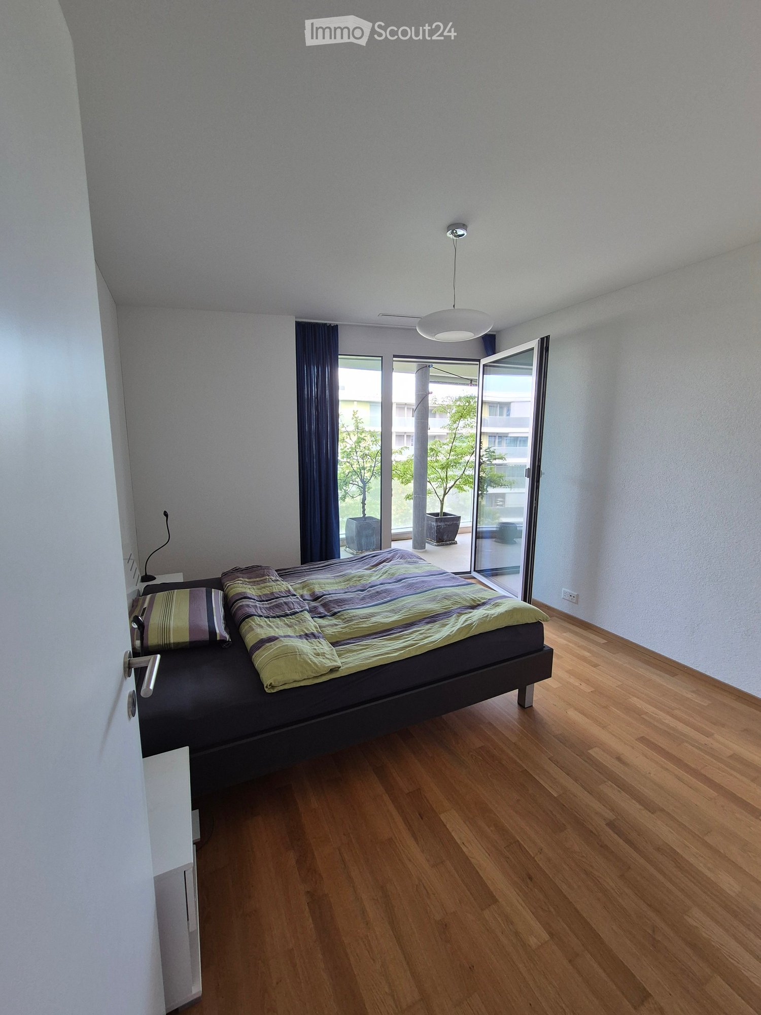 Modern bedroom with wooden floor, large window with dark curtains, bed with striped bedding, pendant light fixture, and a balcony door leading to a small outdoor area with potted plants.