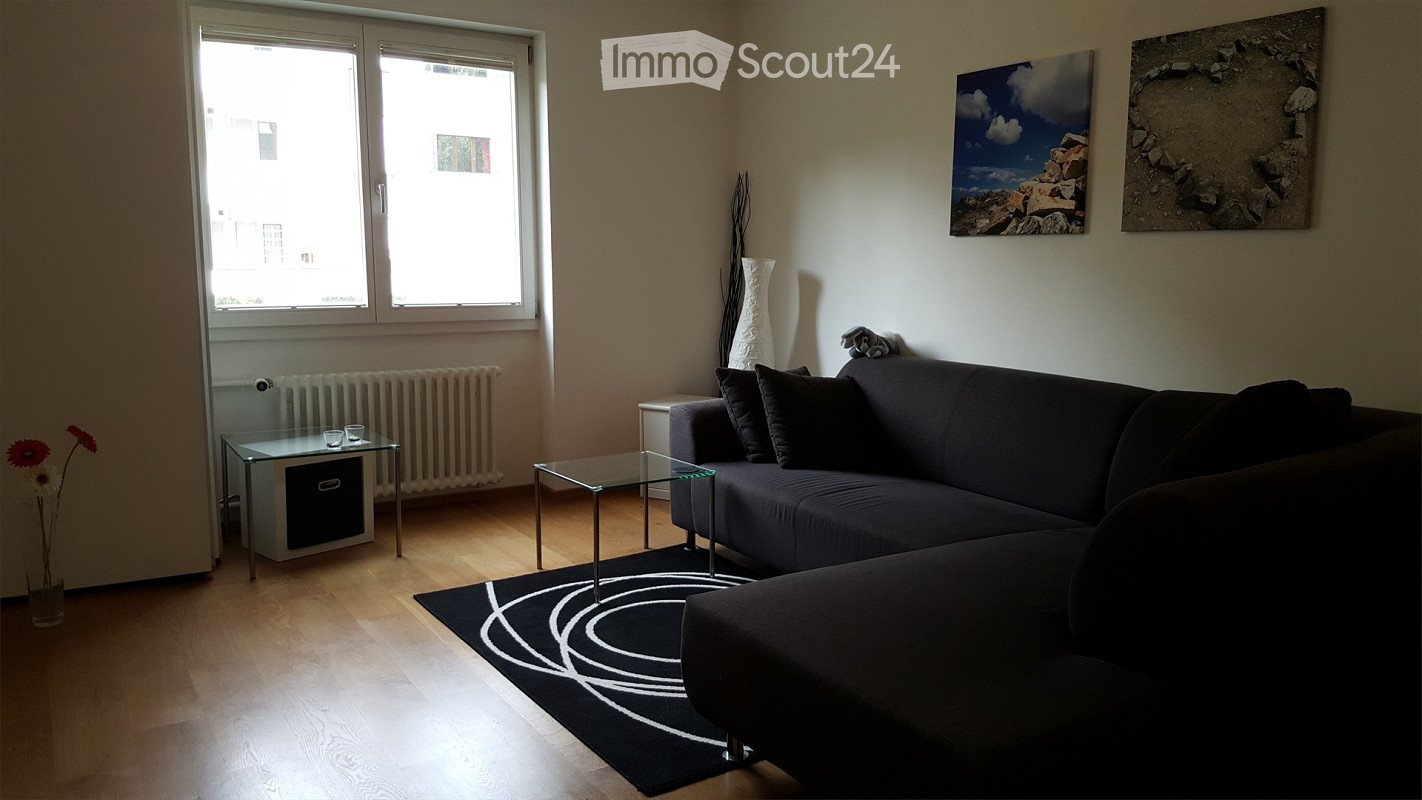 Modern living room with a dark grey sectional sofa, two glass coffee tables, a black and white circular rug, a vase with flowers, and artwork on the walls.