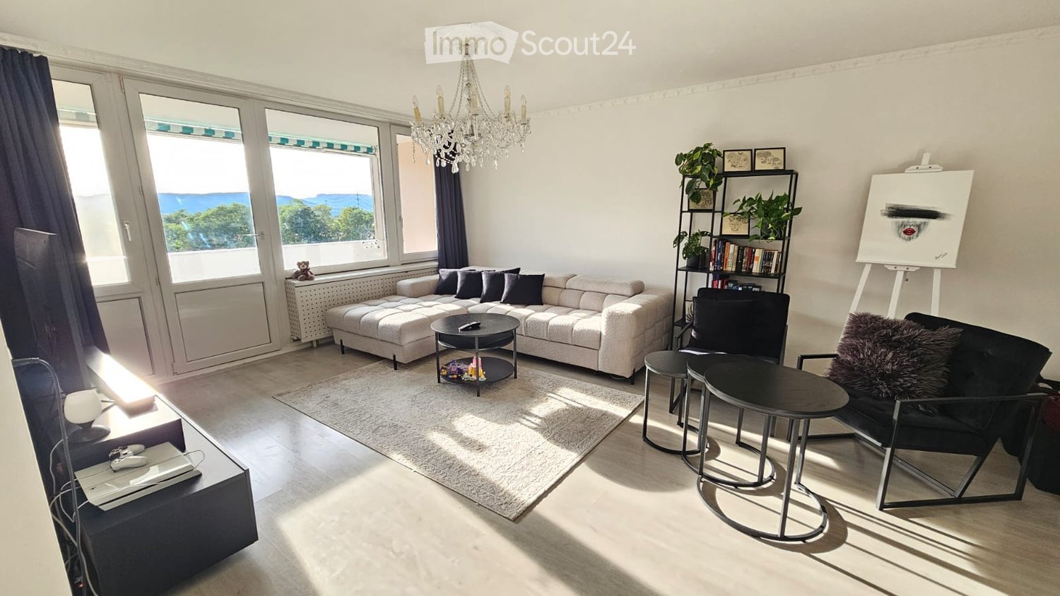 Bright living room with large windows offering a scenic view, featuring a white sectional sofa, coffee table, black chair, small round tables, bookshelf with plants and books, and a chandelier.