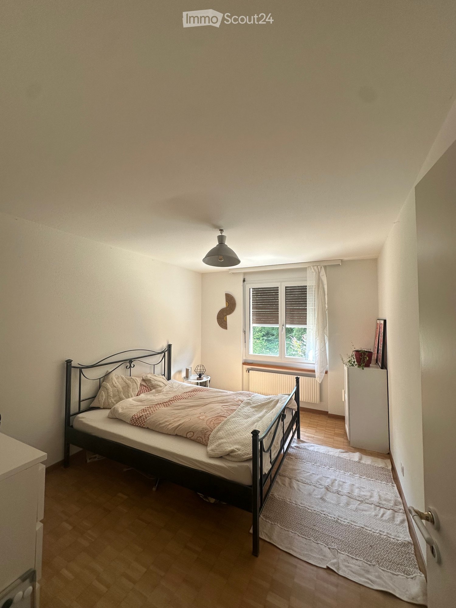 Cozy bedroom with wooden flooring, a metal-framed bed with patterned bedding, a nightstand with a lamp, a window with white curtains, and a small plant on a cabinet.