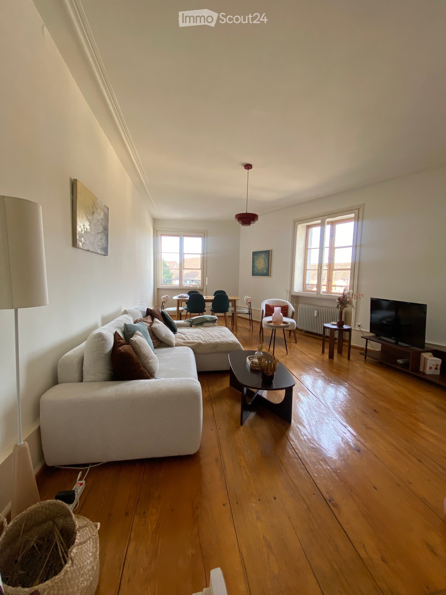 Living room with wooden flooring, large windows, a sectional sofa, coffee table, dining area with chairs and table, television, and decorative items.