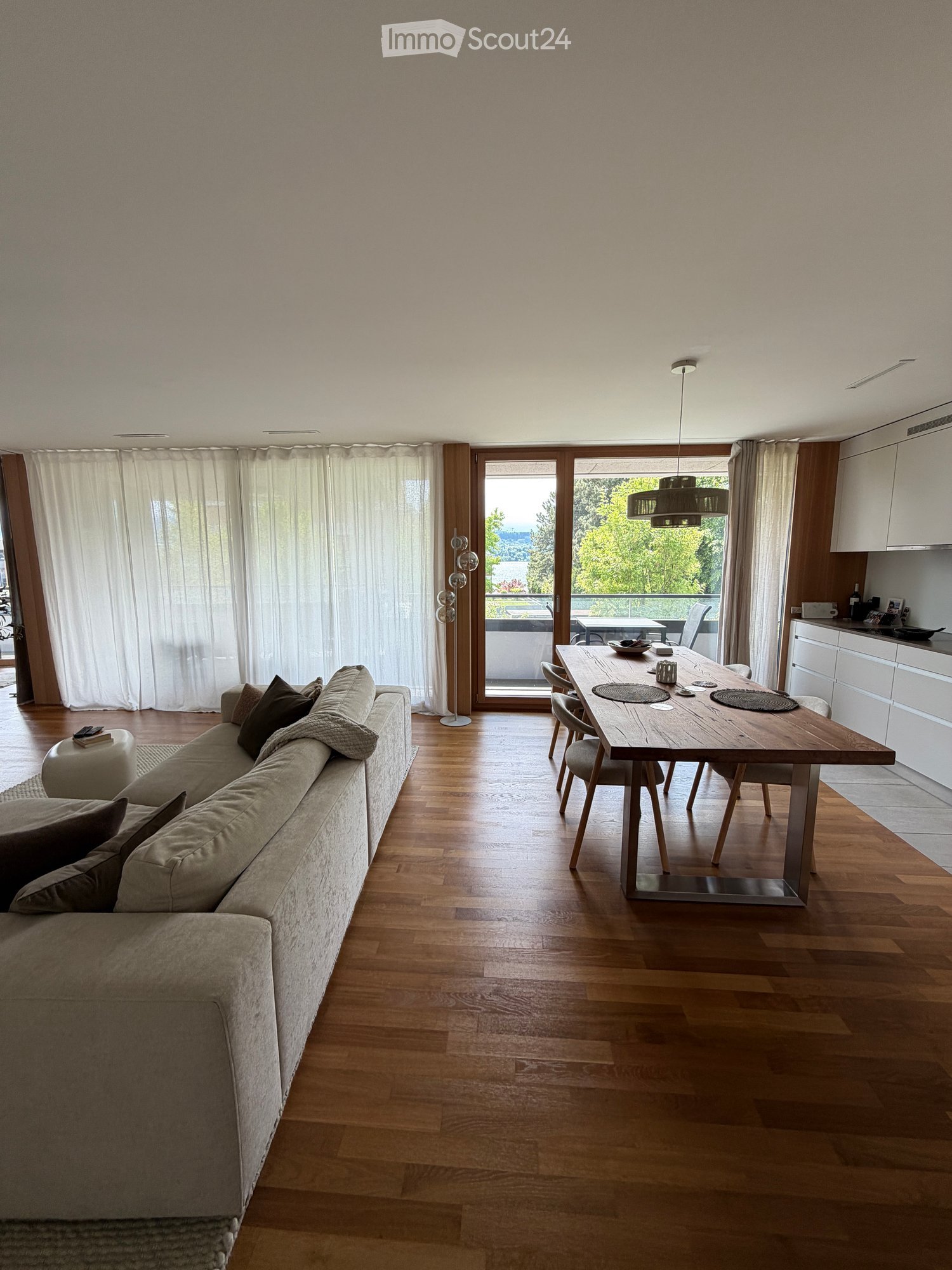 Open-plan living and dining area with wooden flooring, a grey sofa, a wooden dining table with chairs, and a modern kitchen area with white cabinets.