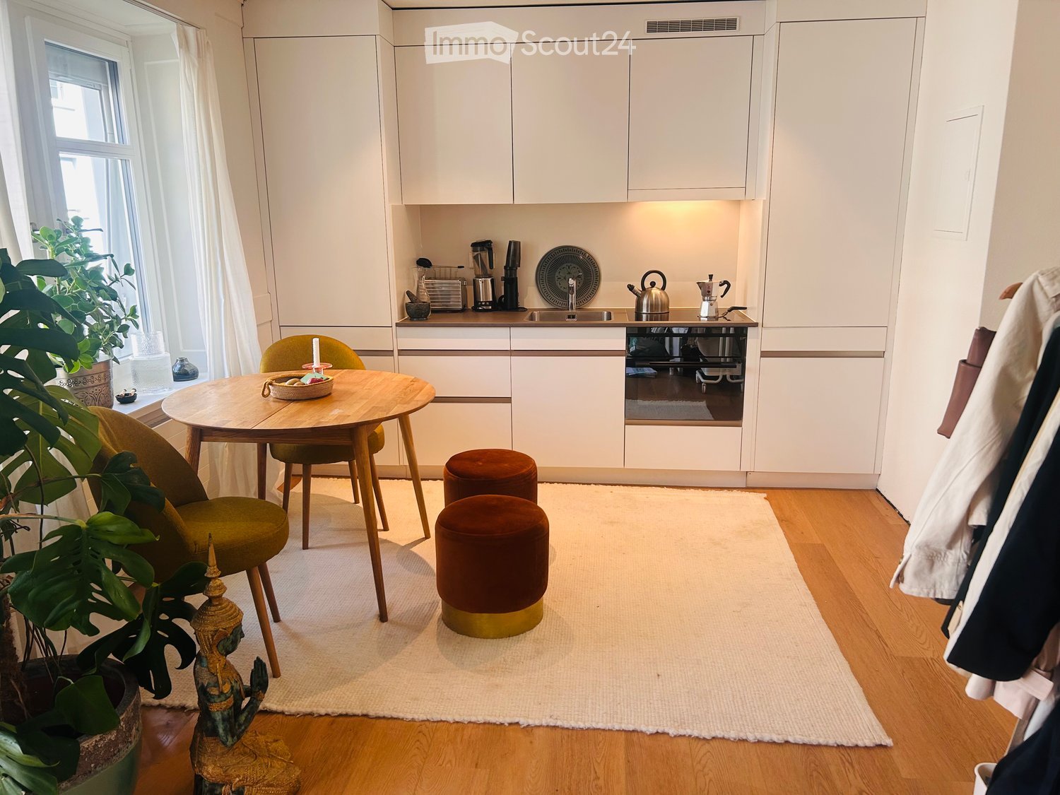 A modern, well-lit kitchen and dining area with white cabinets, a round wooden dining table, two green chairs, two brown stools, a potted plant, and a decorative statue. The kitchen features a sink, teapot, coffee maker, and other small appliances.