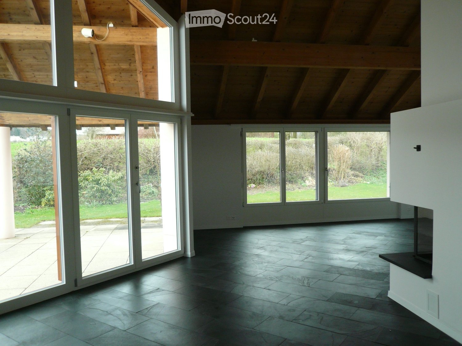 Bright, open-plan room with large glass doors leading to a garden, dark tiled floor, and exposed wooden beams on the ceiling.