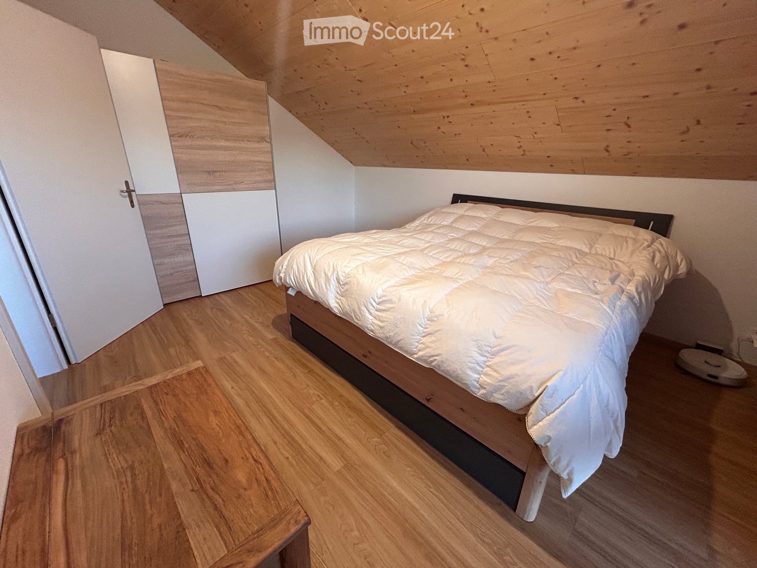 Cozy bedroom with wooden flooring, slanted wooden ceiling, a neatly made bed with white bedding, and a white wardrobe with mirrored doors.