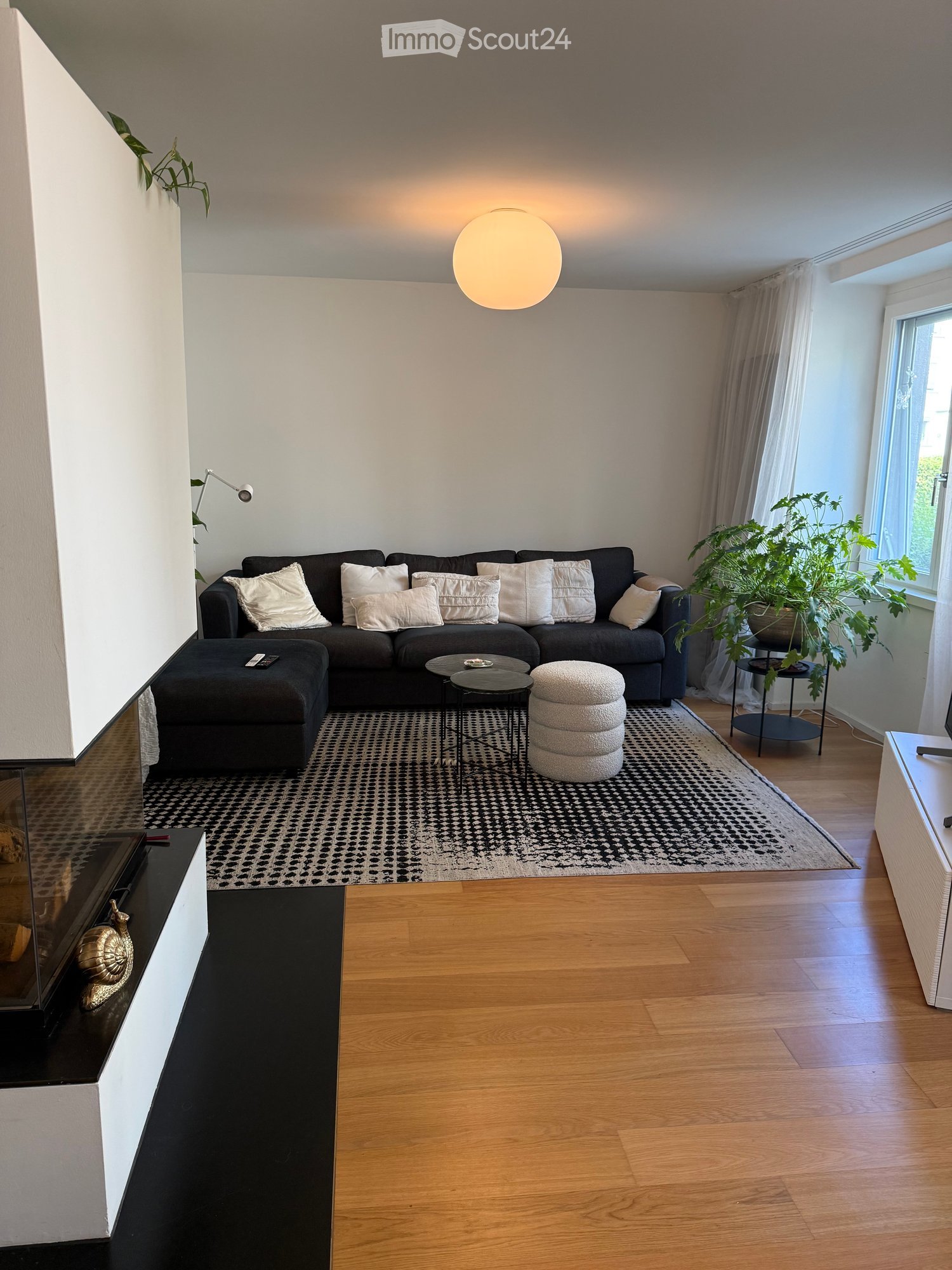 Modern living room with a dark grey sectional sofa, white throw pillows, a black ottoman, a round coffee table, a white round stool, a patterned rug, a large plant, and a fireplace with decorative items.