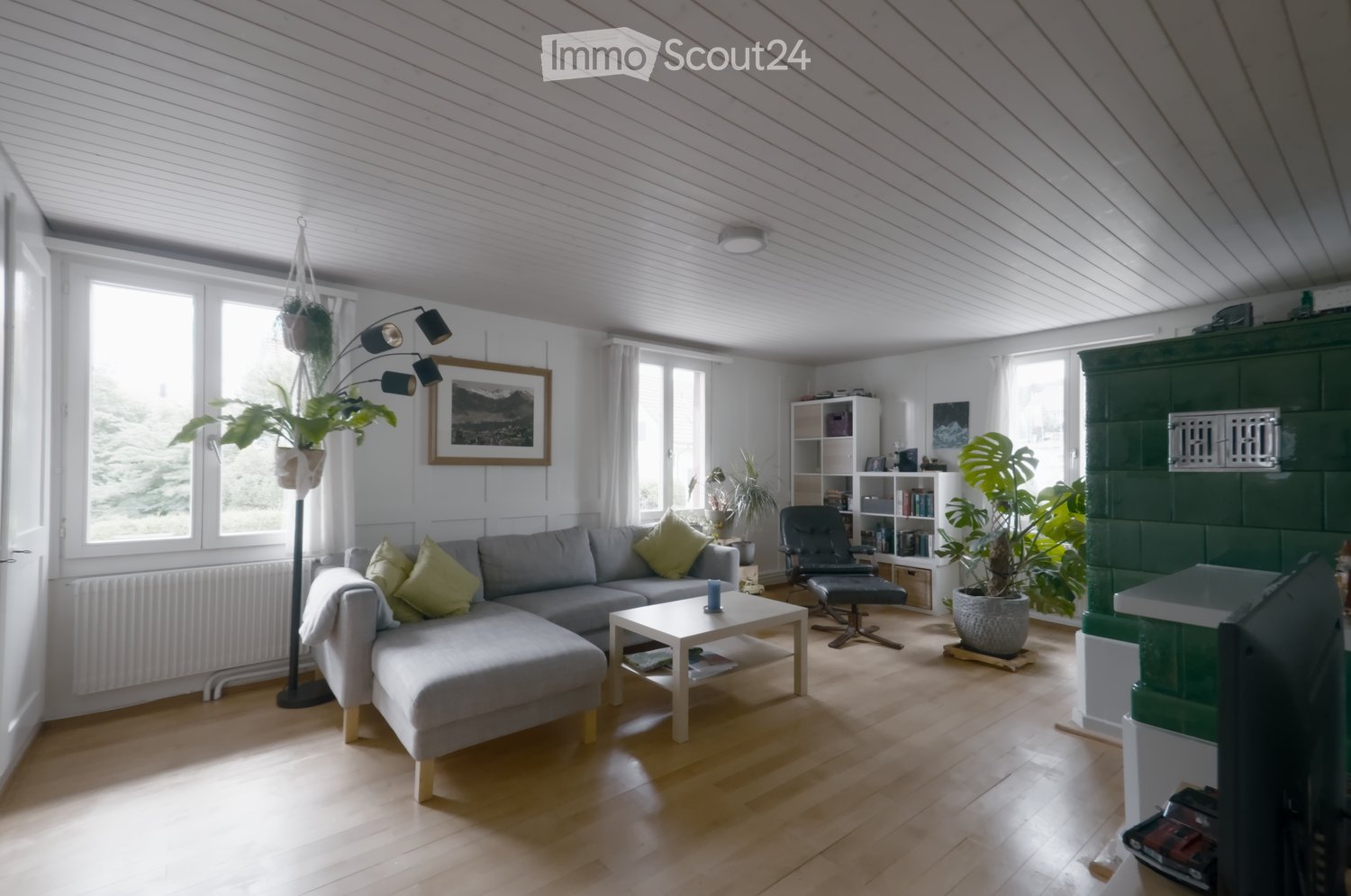 Bright living room with wooden flooring, grey sectional sofa, coffee table, armchair, bookshelf, floor lamp, and potted plants.
