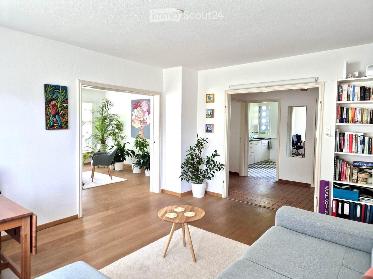 Bright living room with wooden flooring, a grey sofa, a small coffee table, a bookshelf filled with books, and several potted plants. The room features large windows and an open layout connecting to a kitchen area.