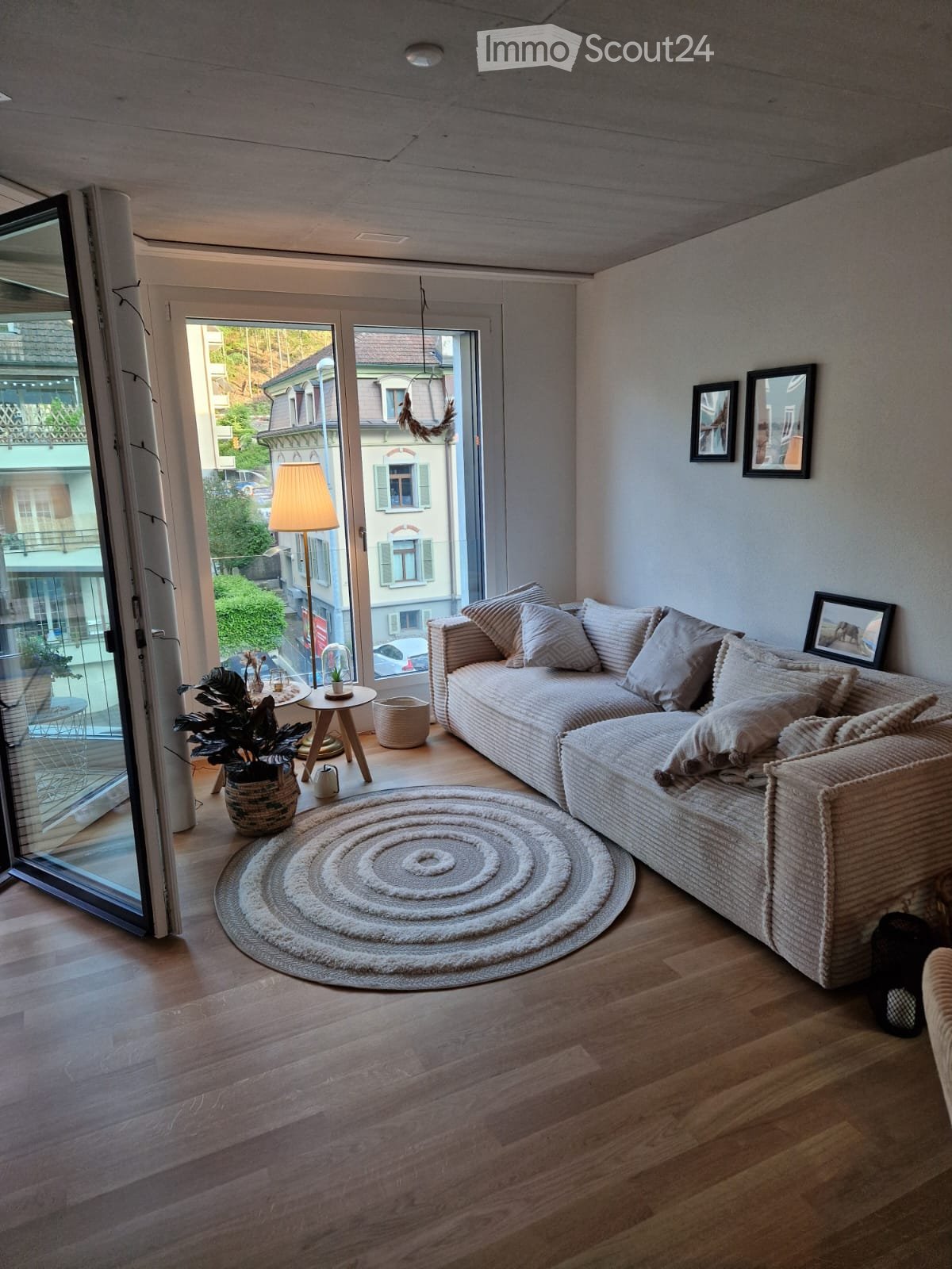 Modern living room with a large sectional sofa, round wooden coffee table, floor lamp, round woven rug, and framed artwork on the wall. The room features wooden flooring and large windows offering a view of the street.