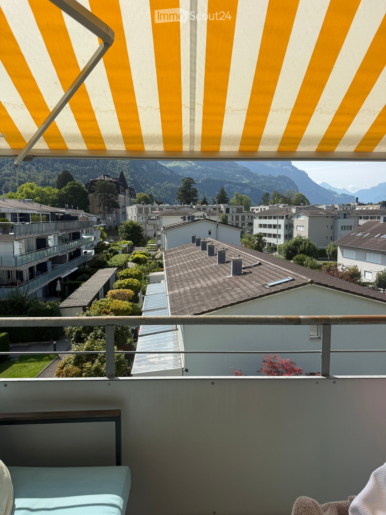Balcony with a view of a town and mountains, featuring a yellow and white striped awning and a metal railing.