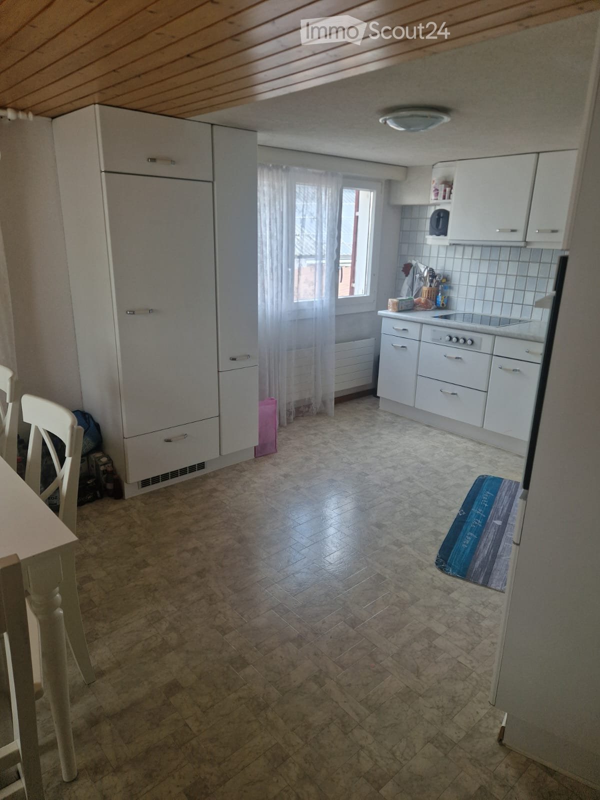Modern kitchen with white cabinets, a refrigerator, an oven, a sink, and a dining area with a table and chairs. The floor is tiled, and there is a window with white curtains.