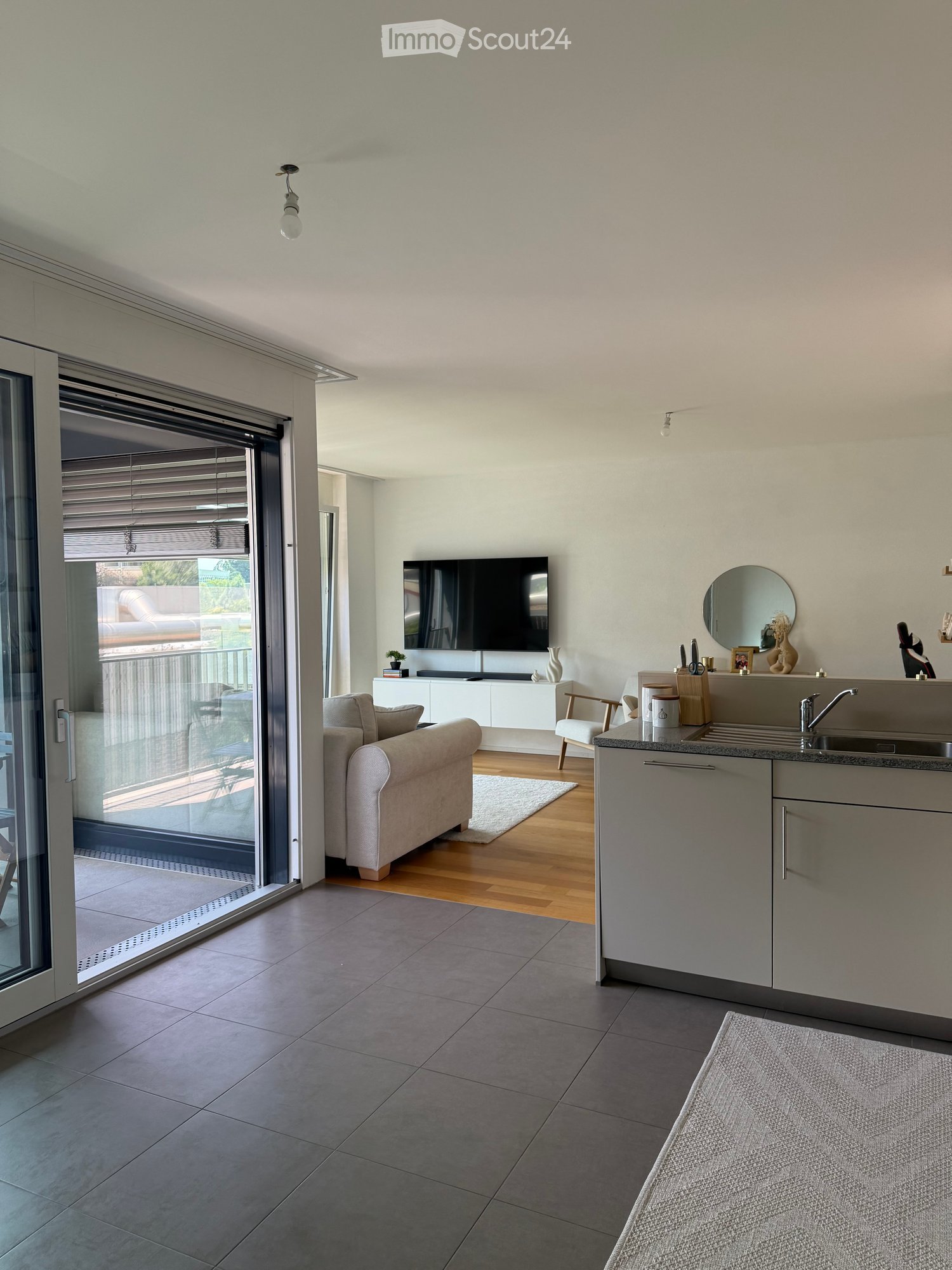 Modern living room with a sofa, chair, television, sink, and a view to a balcony. The room features a mix of wooden and tiled flooring, a white cabinet, and a small table with a vase.
