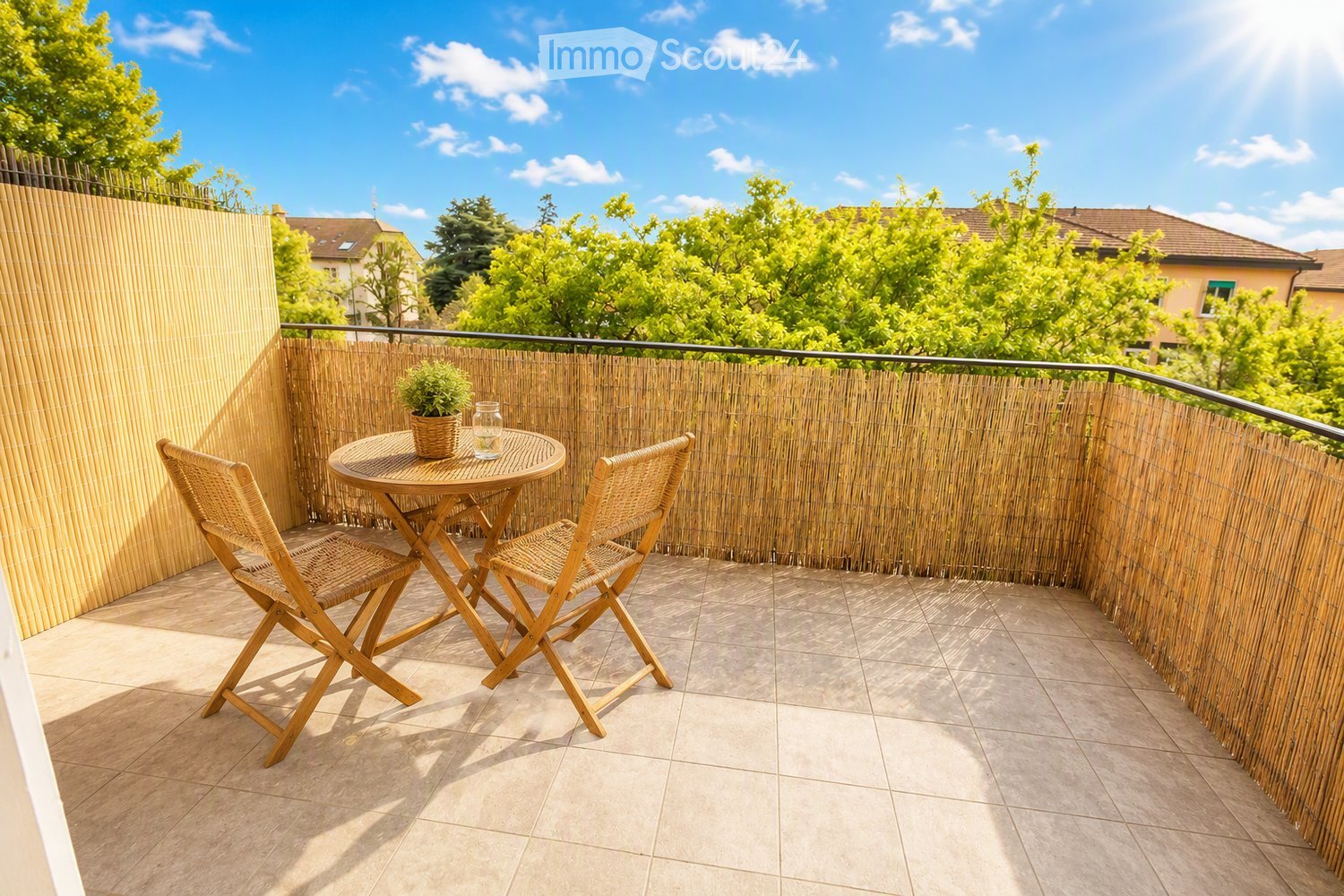 Balcony with a small round table, two wooden chairs, a potted plant, and a glass vase, surrounded by a bamboo fence and overlooking a green area.