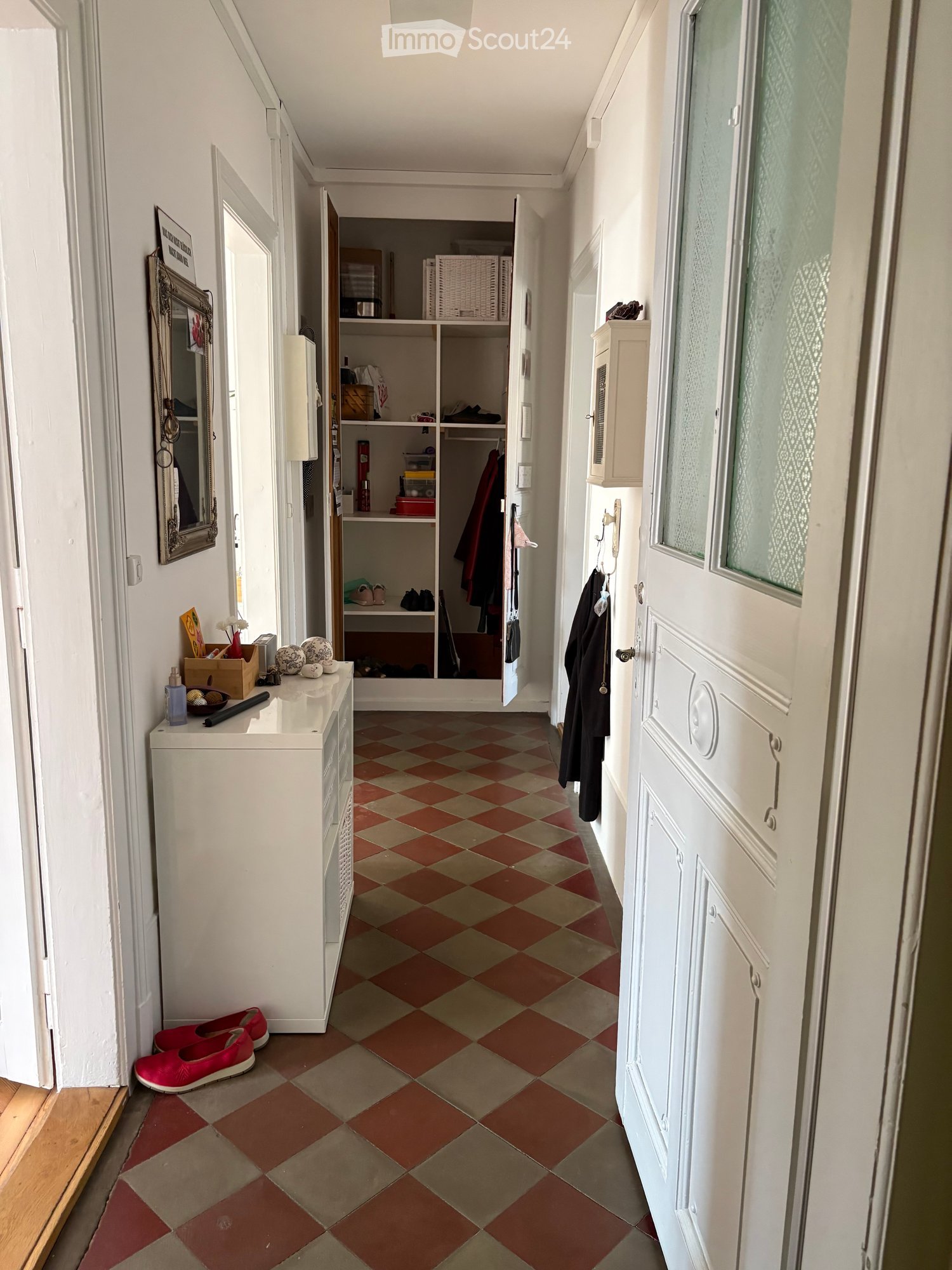 Hallway with checkered tile floor, white walls, open closet with clothes and storage items, small table with decorative items, and a coat rack with jackets.