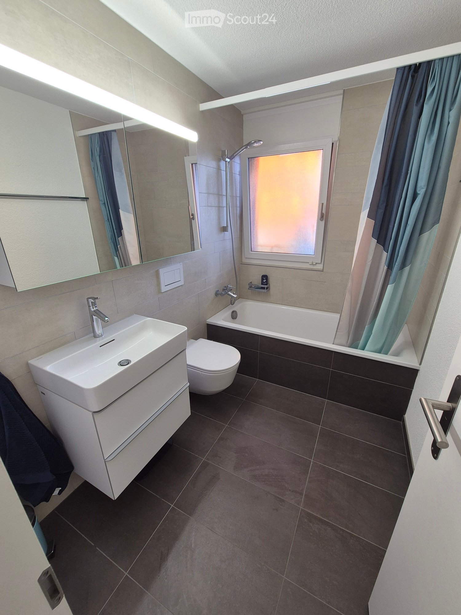 Modern bathroom with a white sink, toilet, bathtub, and large mirror. Features dark tiled flooring and a frosted window with a shower curtain.