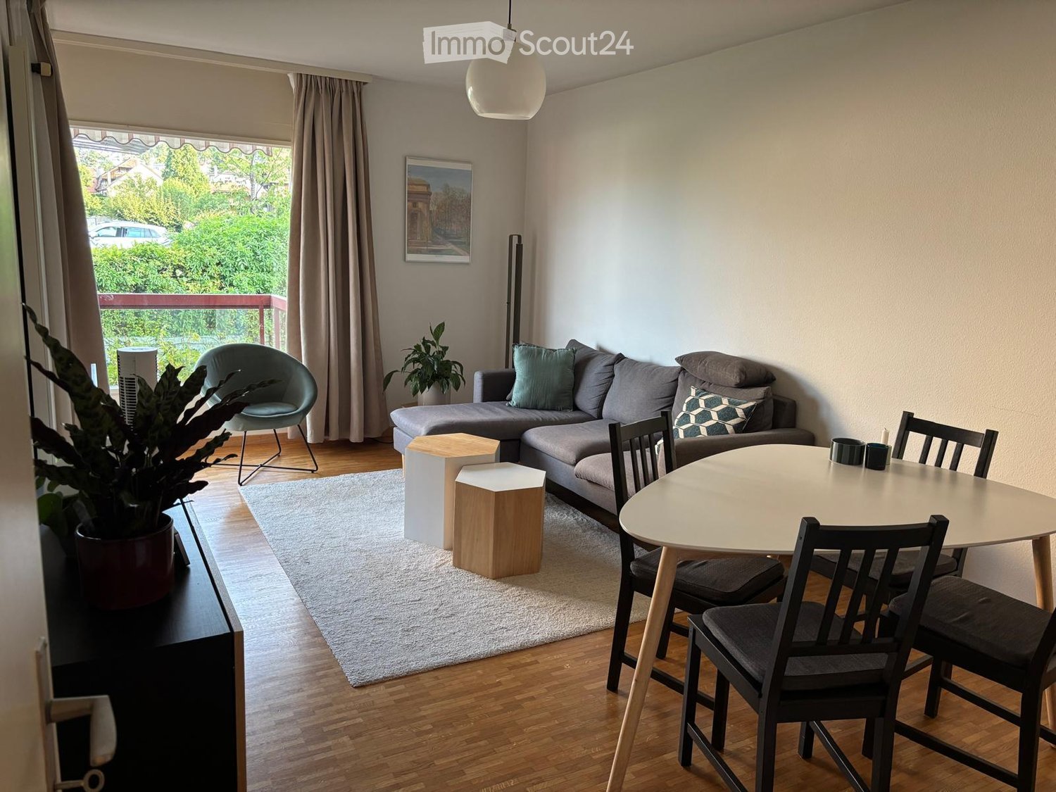 Modern living and dining area with a grey sectional sofa, wooden dining table with chairs, small wooden stools, a potted plant, and a picture frame on the wall.