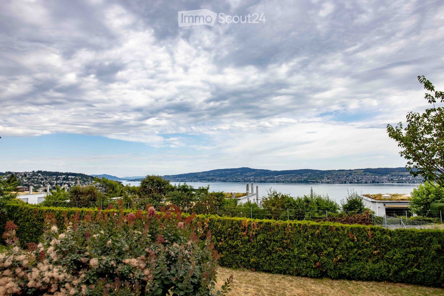 A well-maintained garden with neatly trimmed hedges and flowering shrubs, overlooking a scenic waterfront with distant hills and a cityscape.