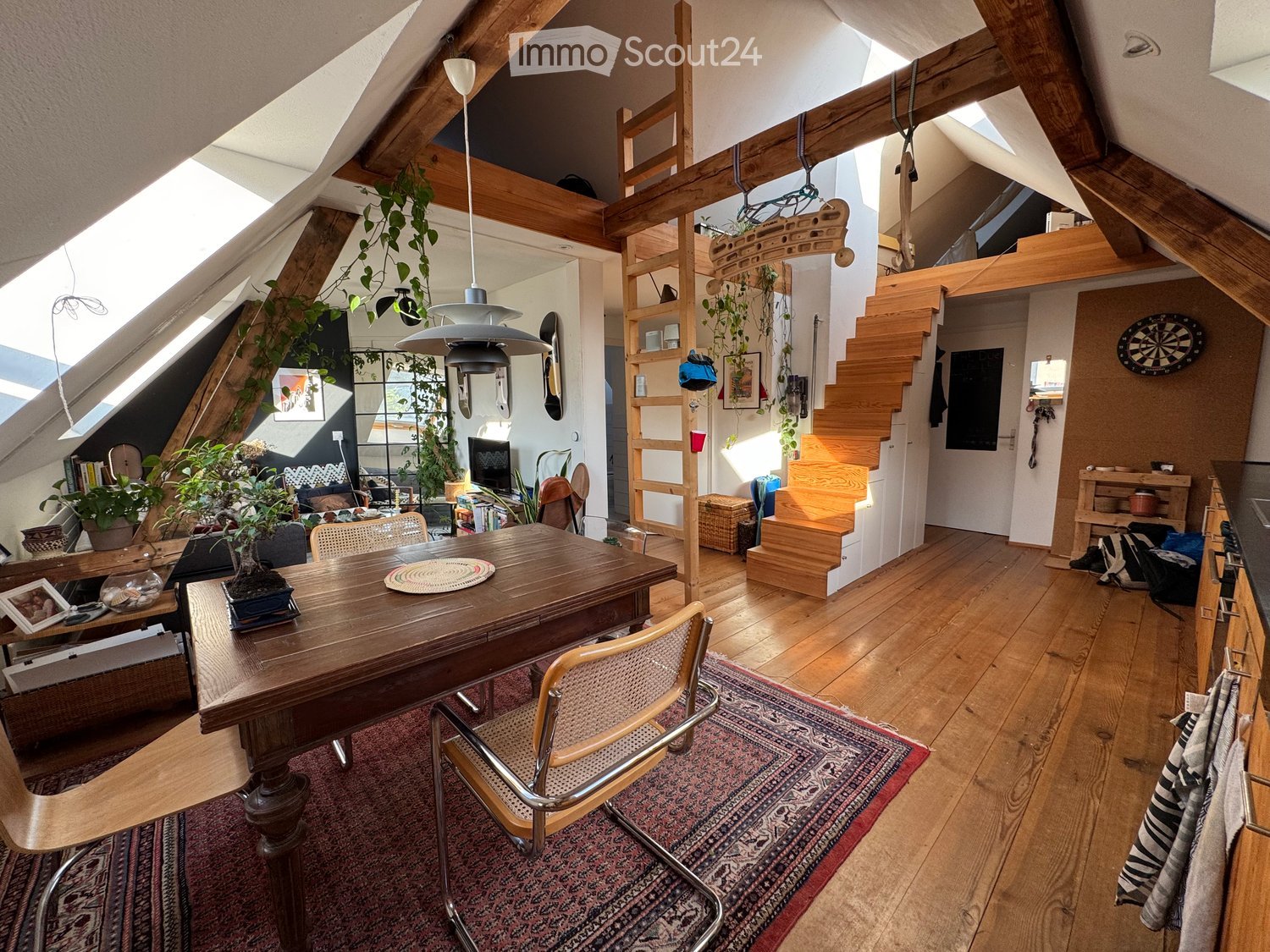 Cozy attic space with wooden beams, skylights, and exposed wooden floors. Features a dining table with chairs, pendant light, shelves, staircase leading to a loft area, and various decorative items including plants and artwork.