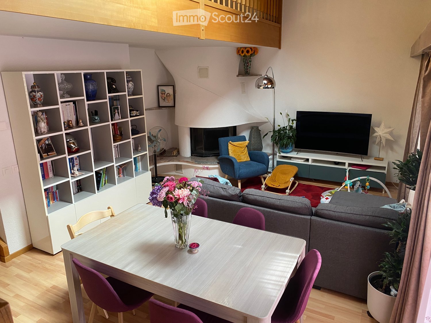 Modern living room with a fireplace, grey sofa, blue armchair, dining table with chairs, shelves, TV, and decorative items.