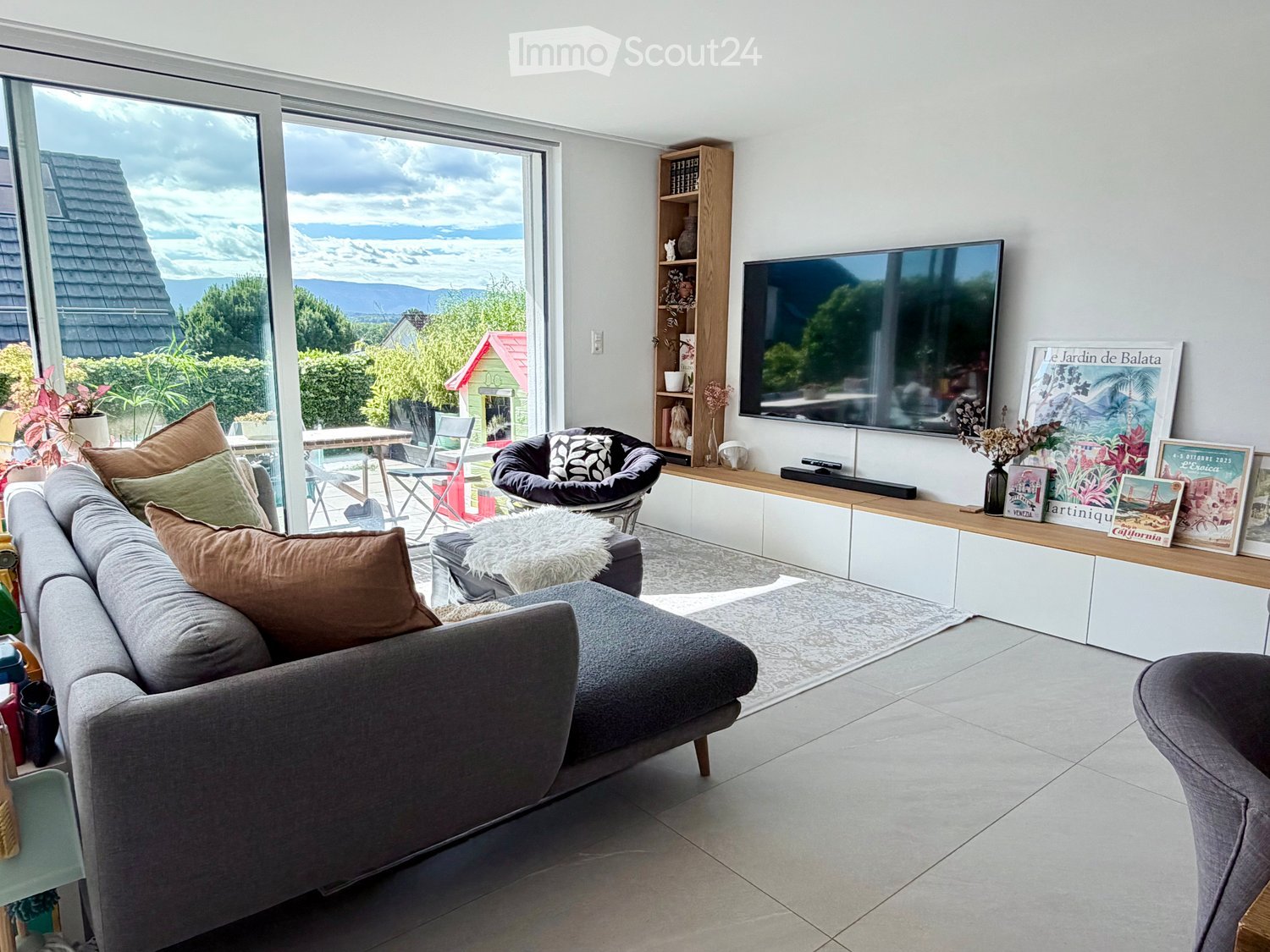 Modern living room with large windows offering a scenic view, a grey sectional sofa, a black egg chair, a wooden shelf, and a flat-screen TV.