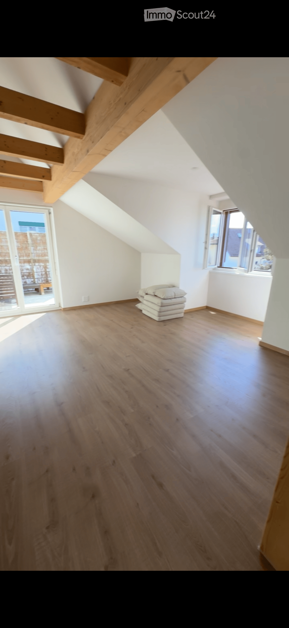 Empty attic room with wooden floor, white walls, exposed wooden beams, and large windows letting in natural light.