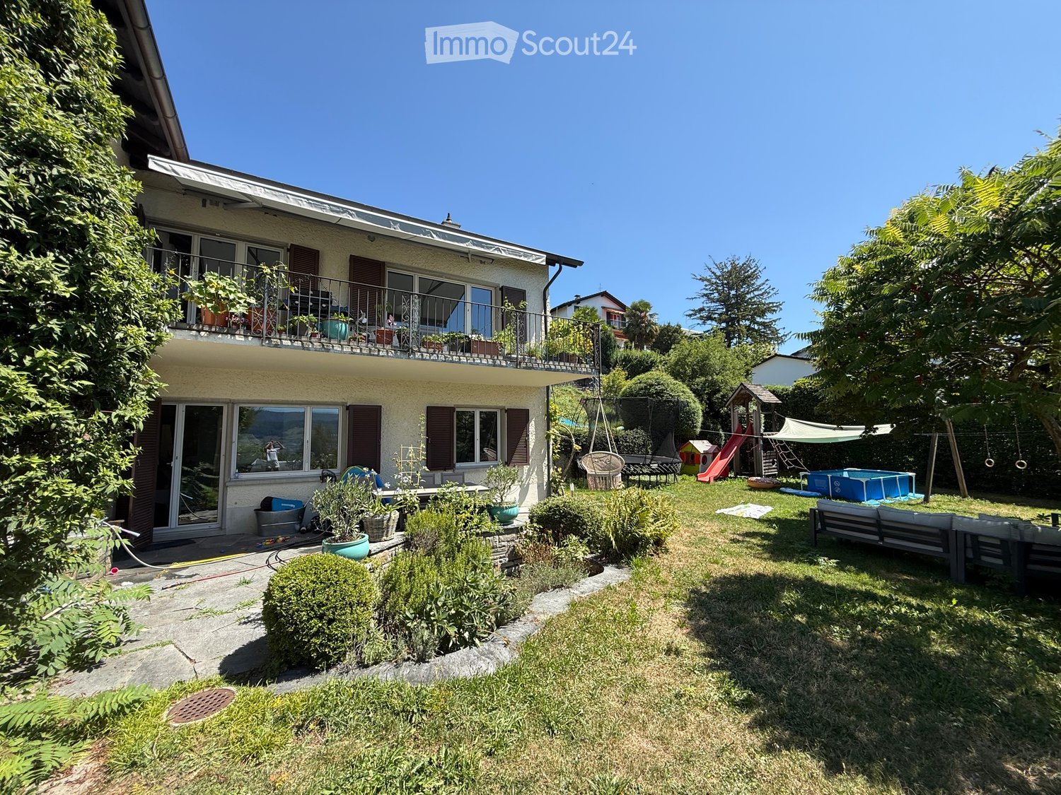 Garden area with a swing, slide, and small pool, surrounded by a two-story house with a balcony and large windows.