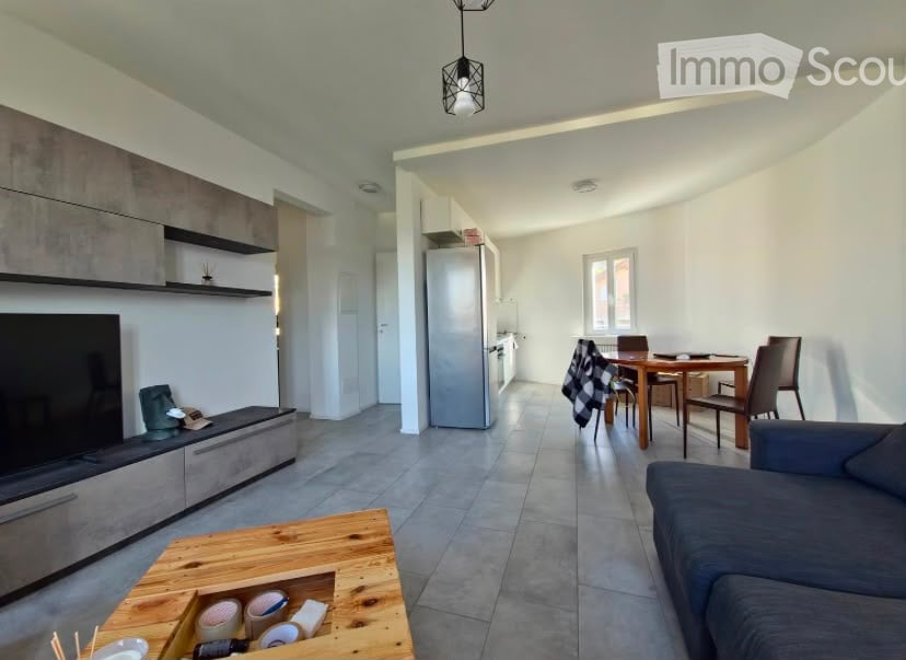 Modern open-plan living and dining area with grey sofa, wooden coffee table, dining table with chairs, and kitchen area featuring cabinets and a refrigerator.